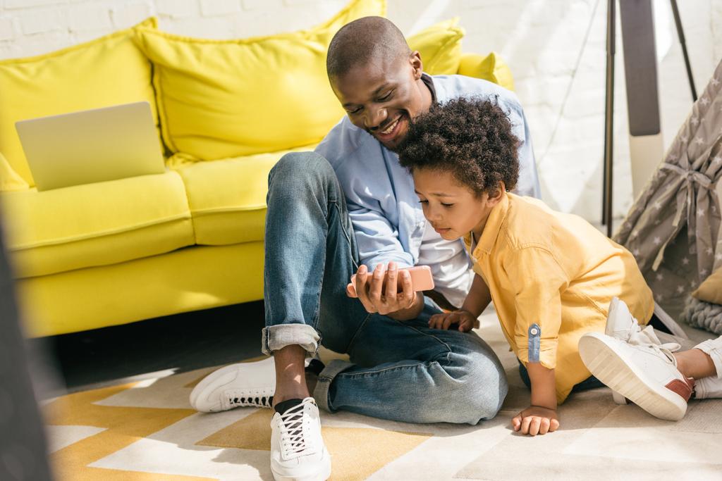 lachende Afro-Amerikaanse en weinig zoon smartphone samen thuis gebruiken - Foto, afbeelding
