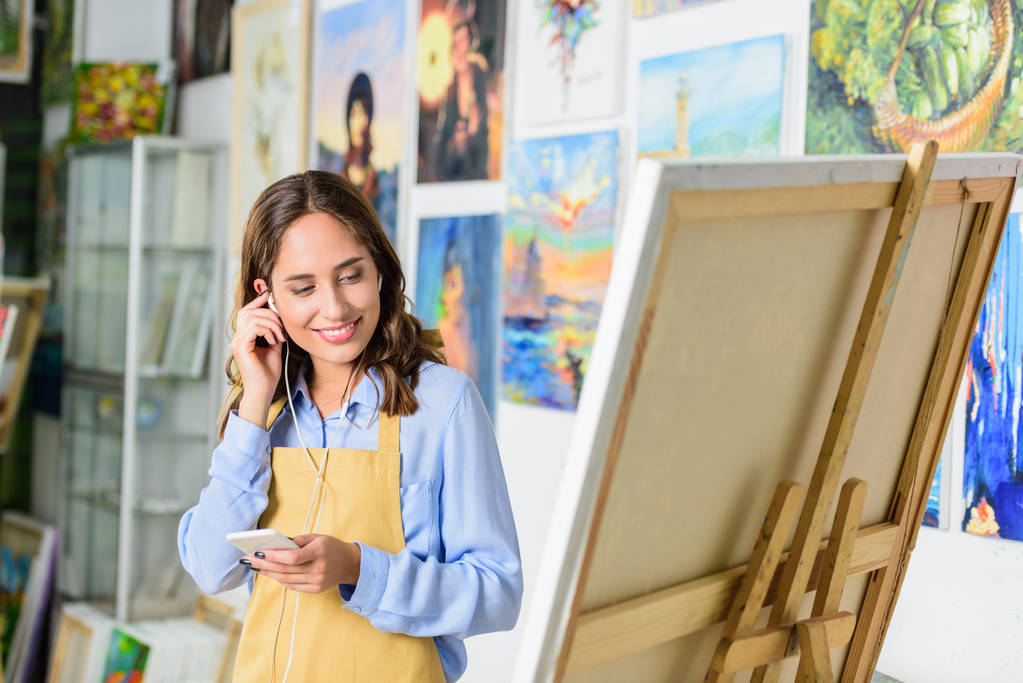 prachtige vrouwelijke artiest luisteren naar muziek met smartphone en kijken naar doek in werkplaats - Foto, afbeelding