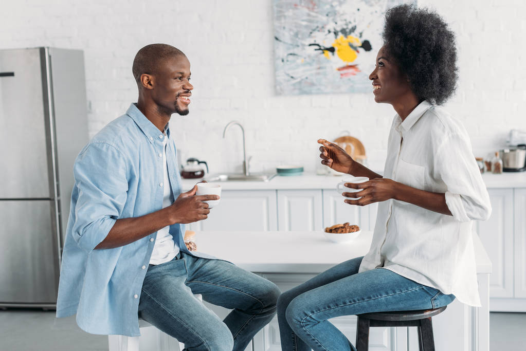 Zijaanzicht van Afrikaanse Amerikaanse echtpaar met kopjes koffie in de ochtend samen in de keuken thuis - Foto, afbeelding