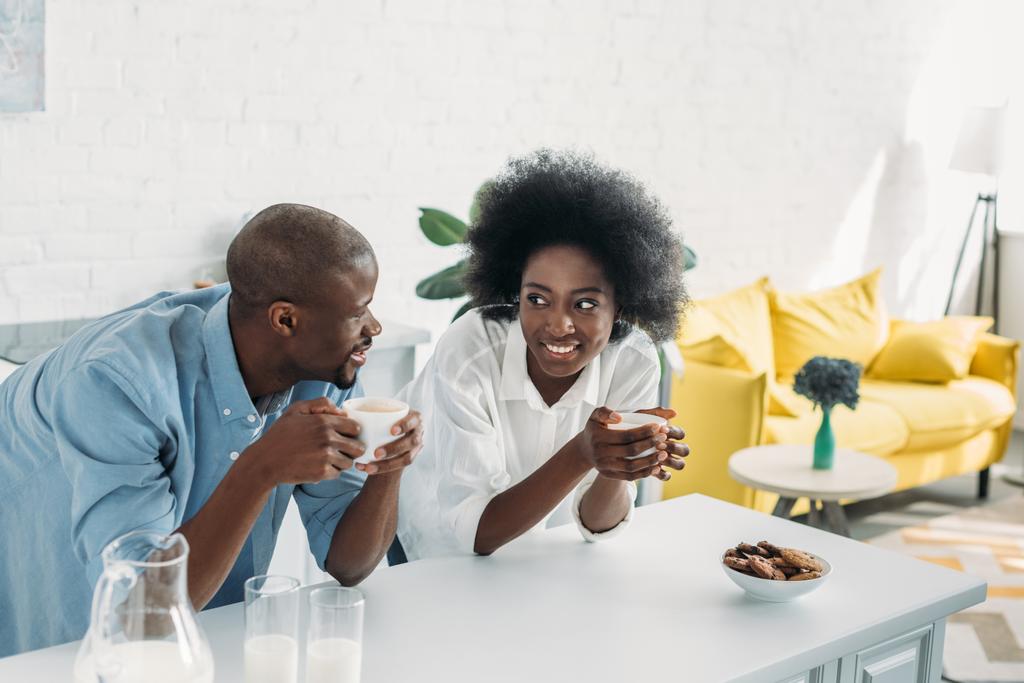 χαμογελώντας αφρικανική αμερικανική ζευγάρι με φλιτζάνια του καφέ πρωί μαζί στην κουζίνα στο σπίτι - Φωτογραφία, εικόνα