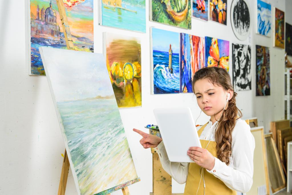 niño serio de pie con la tableta en el taller de la escuela de arte y apuntando sobre lienzo
 - Foto, imagen