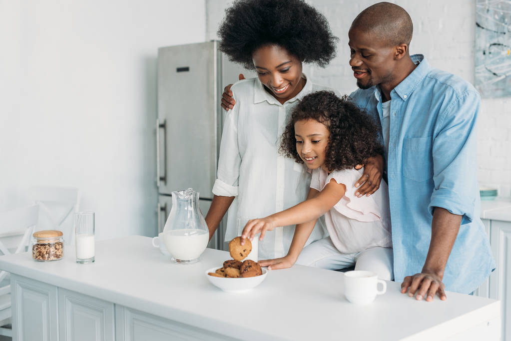 Afrikanisch-amerikanisches Kind nimmt Kekse, während Eltern in der Nähe in der Küche zu Hause stehen - Foto, Bild