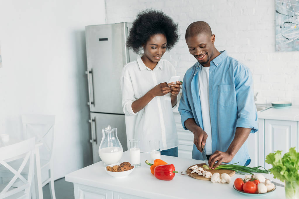 усміхнений афроамериканський чоловік готує сніданок і дружина зі смартфоном, що стоїть поруч на кухні вдома
 - Фото, зображення