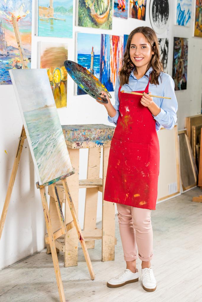 bela artista feminina segurando pincel de pintura e paleta perto de lona em oficina
 - Foto, Imagem