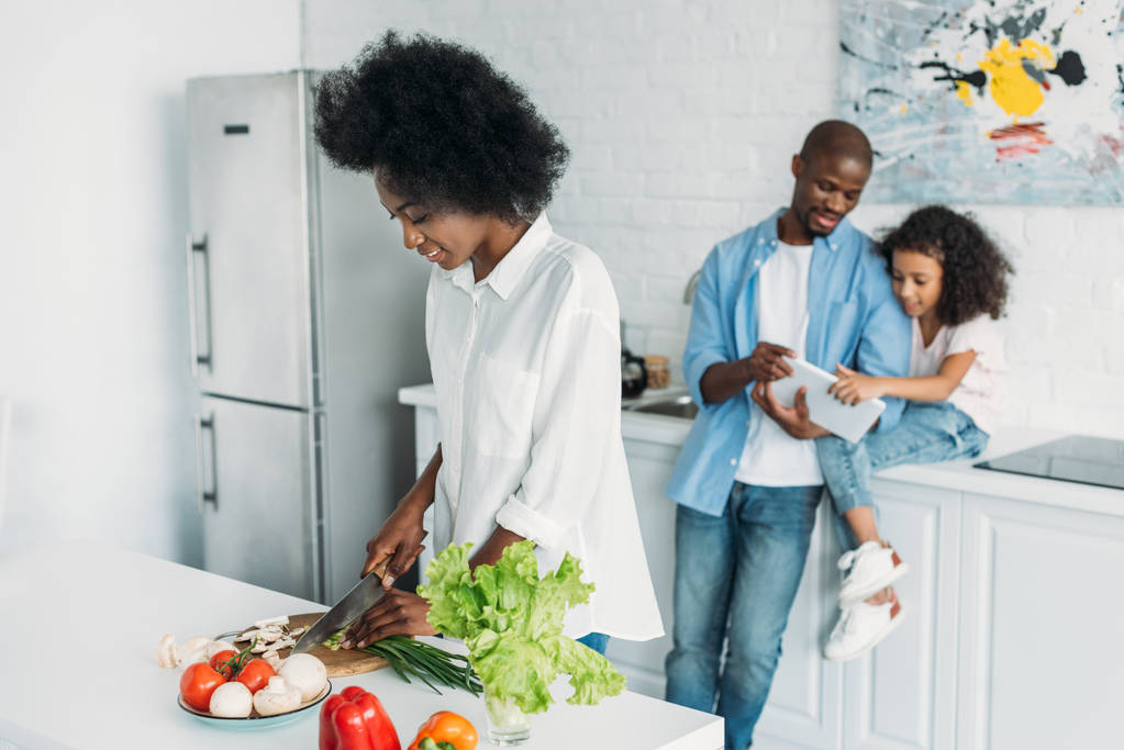 Selektiver Fokus der afrikanisch-amerikanischen Frau beim Kochen des Frühstücks mit der Familie mit Tablette hinter der Theke in der heimischen Küche - Foto, Bild