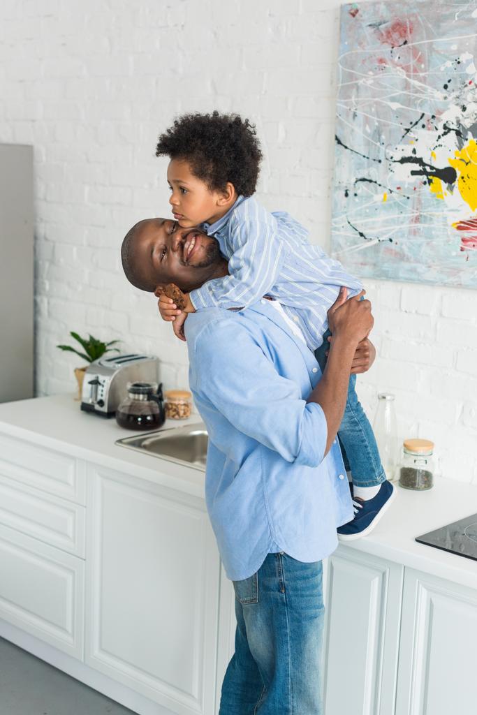african american man holding little son with cookie in kitchen at home - Photo, Image