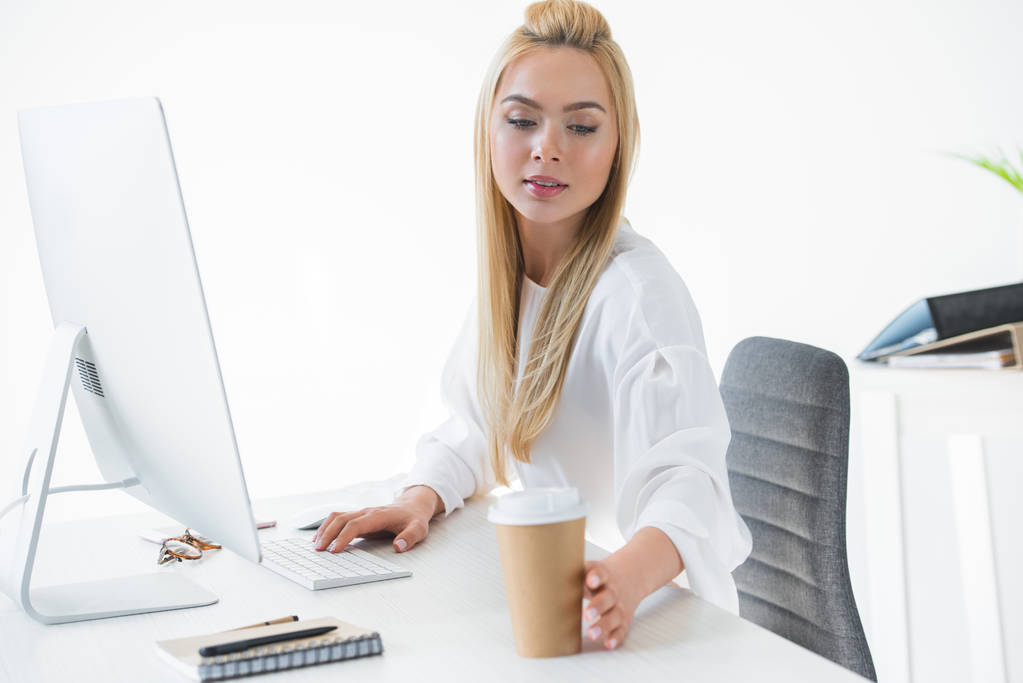 красивая молодая деловая женщина тянется за бумажной чашкой с кофе, используя настольный компьютер на рабочем месте
 - Фото, изображение