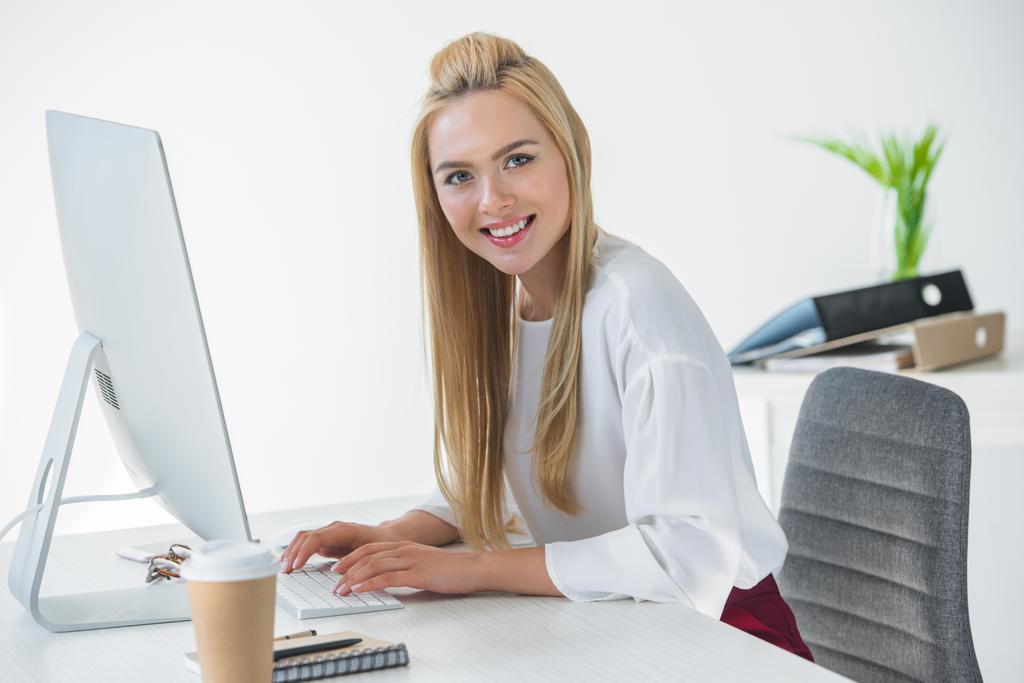 krásná mladá podnikatelka se usmívá na kameru při používání stolní počítač v kanceláři - Fotografie, Obrázek