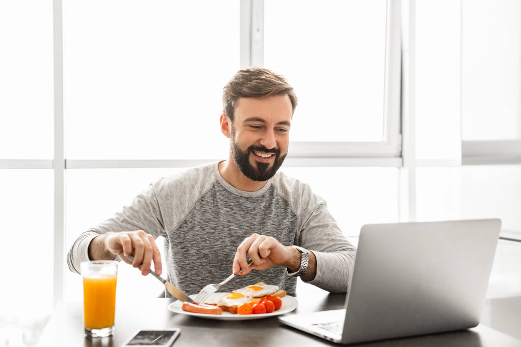 Αξύριστο ικανοποιημένος άνθρωπος 30s φορώντας ρούχα τρώει τηγανητό  - Φωτογραφία, εικόνα