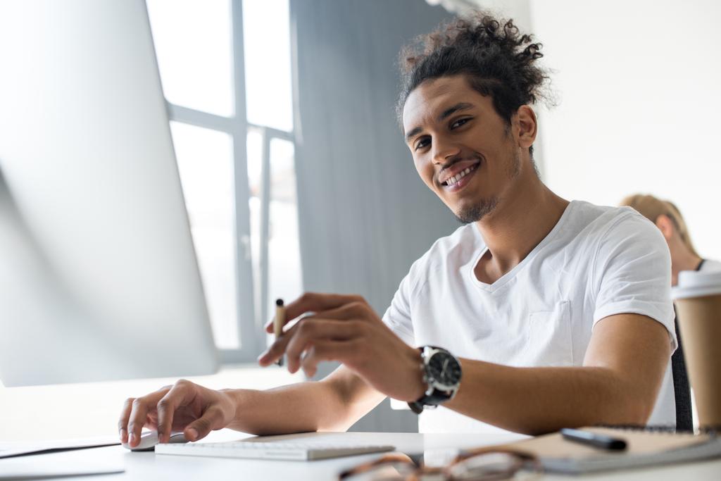 yakışıklı genç Afro-Amerikan adam Office masaüstü bilgisayarla çalışırken kameraya gülümseyen - Fotoğraf, Görsel