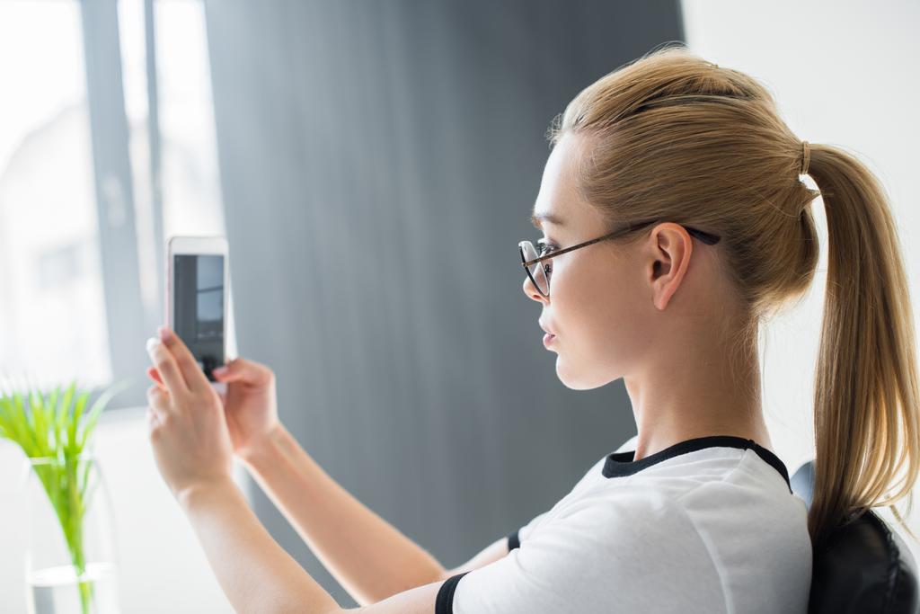 vista lateral da jovem loira tomando selfie com smartphone no escritório
  - Foto, Imagem