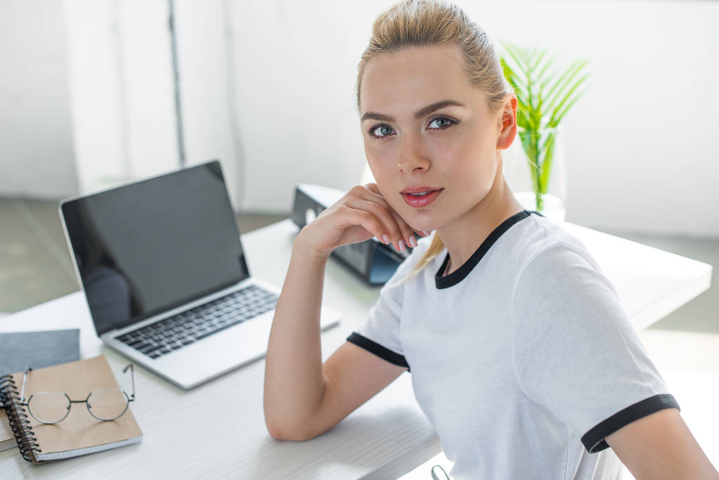 mooie jonge vrouw camera kijken tijdens het werken met de laptop in kantoor - Foto, afbeelding
