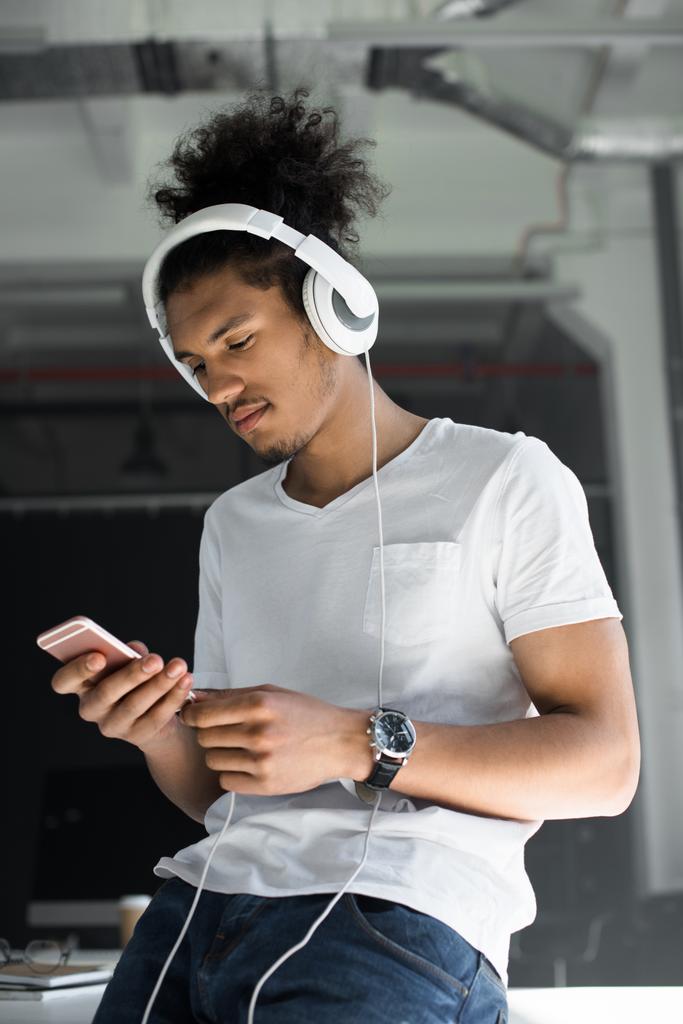 όμορφος νεαρός αφρικανική αμερικανική άνθρωπος στα ακουστικά χρησιμοποιώντας το smartphone στο γραφείο    - Φωτογραφία, εικόνα