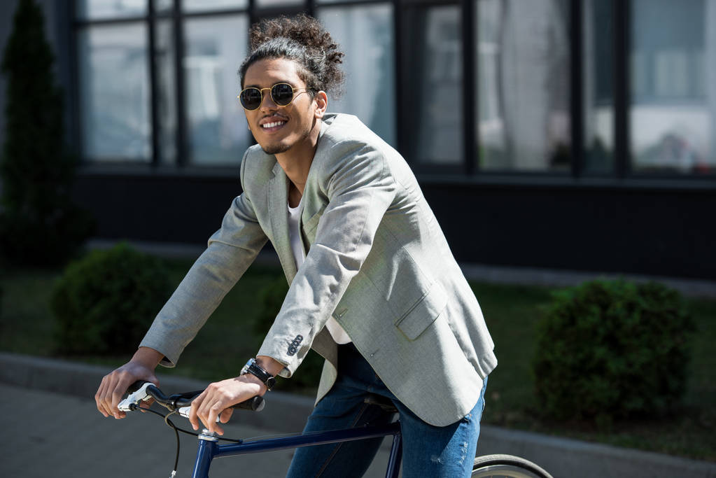 stijlvolle jonge Afro-Amerikaanse man in zonnebril zittend op de fiets en glimlachend op camera - Foto, afbeelding