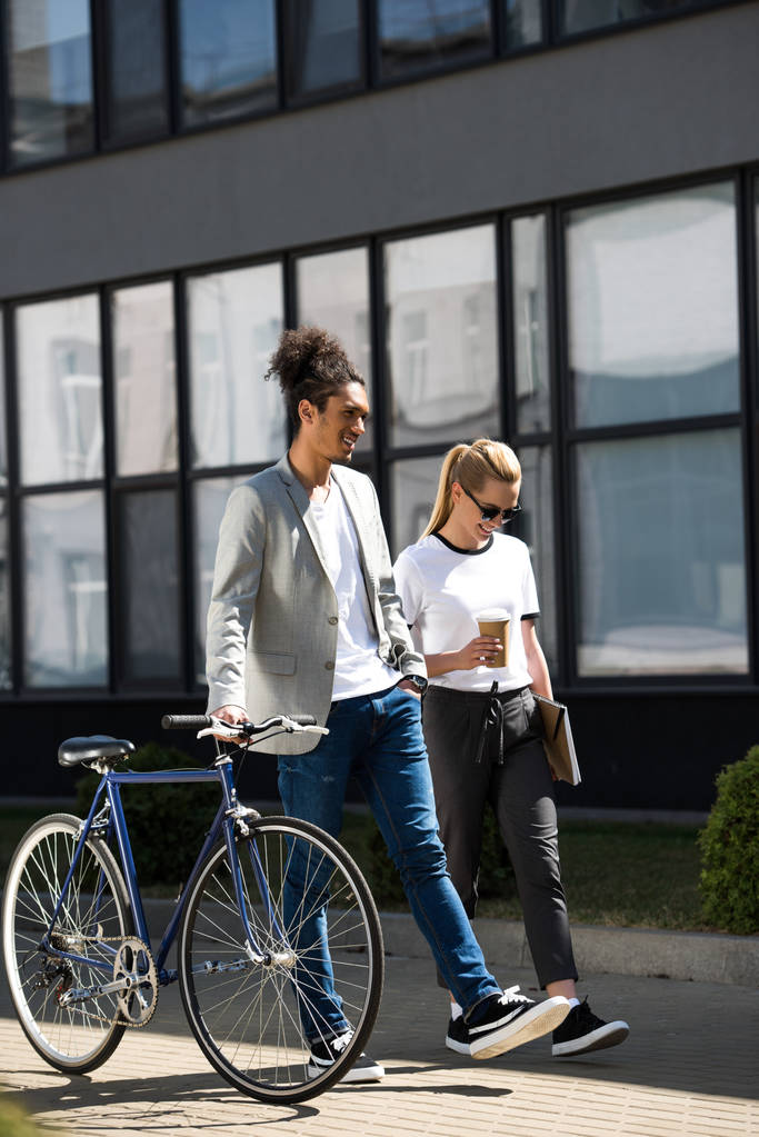 sonriente joven pareja multiétnica con bicicleta caminando por la calle
 - Foto, imagen