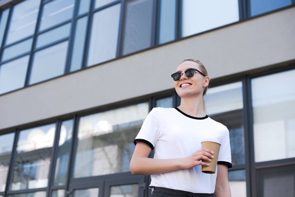 vista de ángulo bajo de hermosa mujer joven sonriente sosteniendo café para ir y mirar hacia otro lado mientras camina por la calle
 - Foto, Imagen