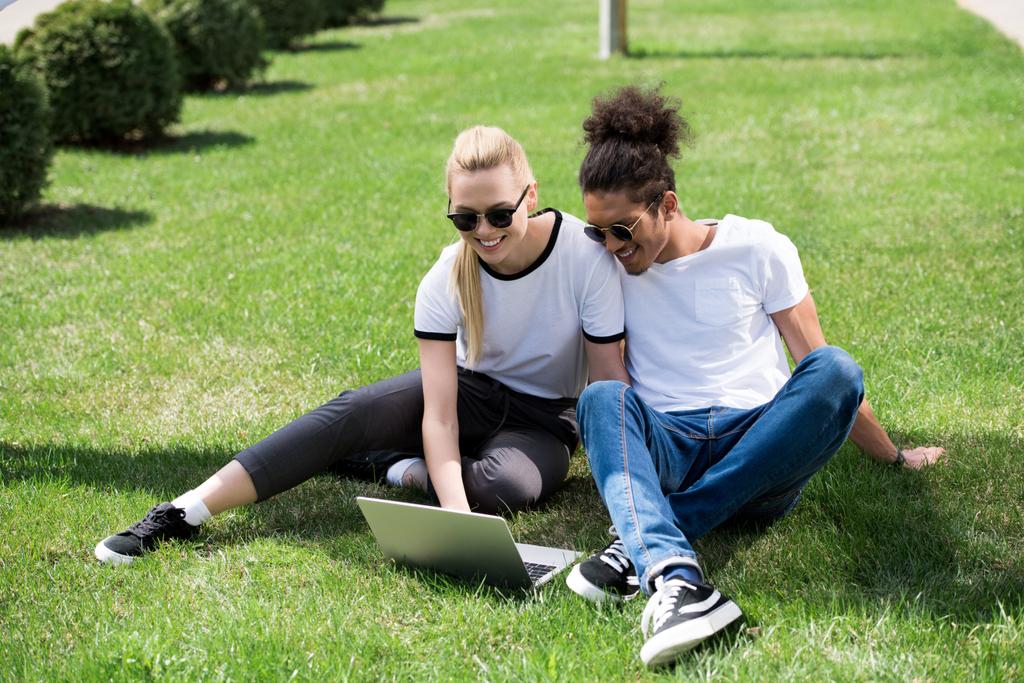 feliz joven pareja multiétnica sentado en la hierba y el uso de ordenador portátil
 - Foto, Imagen