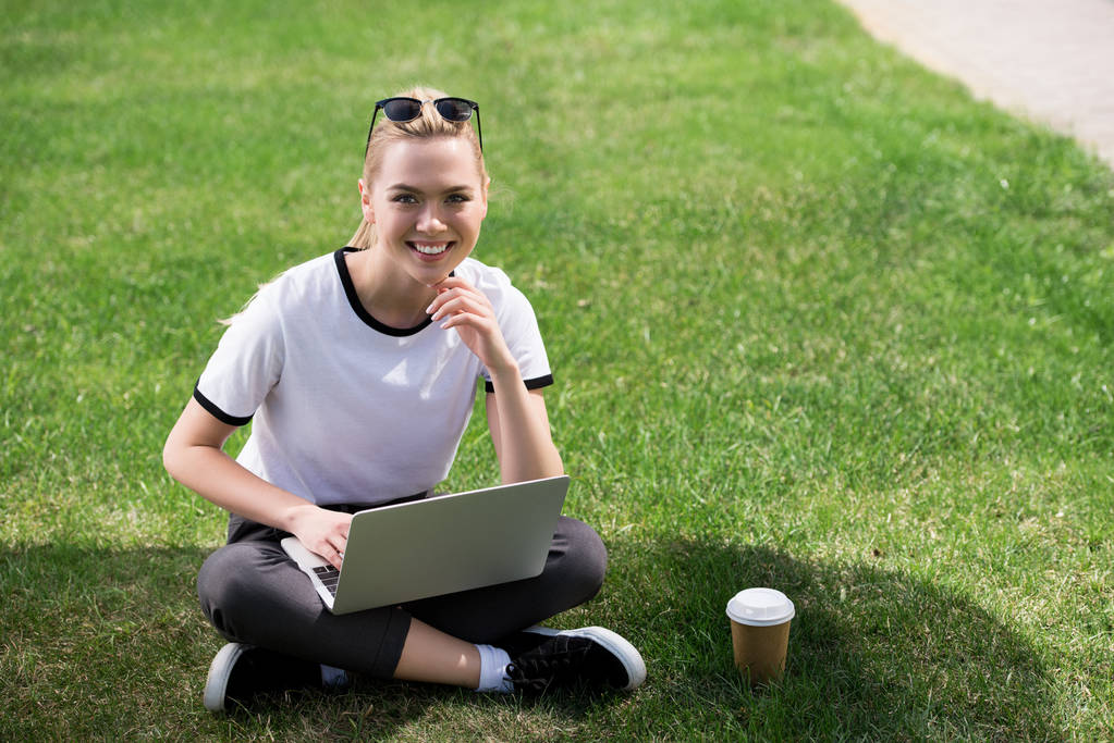 kaunis blondi tyttö hymyilee kameralle, kun käytät kannettavaa tietokonetta ruoholla
 - Valokuva, kuva