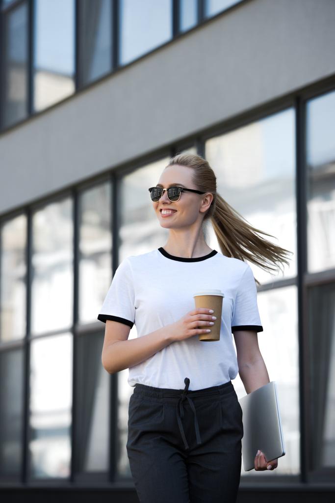 chica rubia sonriente con ordenador portátil y taza de café desechable mirando hacia el exterior edificio de oficinas
 - Foto, Imagen