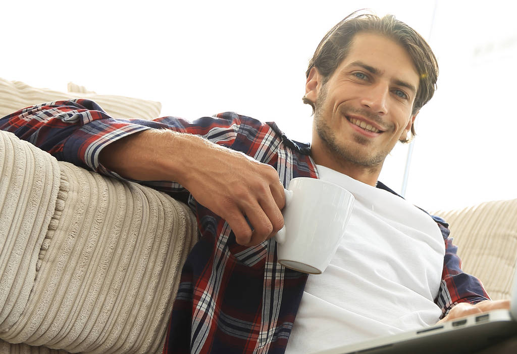 jeune homme avec ordinateur portable tenant une tasse assise sur le sol près du canapé
 - Photo, image