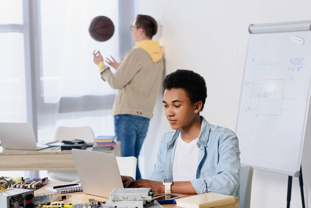 african american teen boy using laptop and friend playing with basketball ball at home - Photo, Image