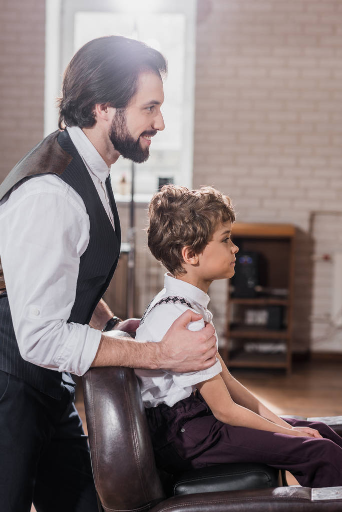 Zijaanzicht van knappe barber kijken spiegelbeeld van kleine jongen zitten in stoel barbershop - Foto, afbeelding