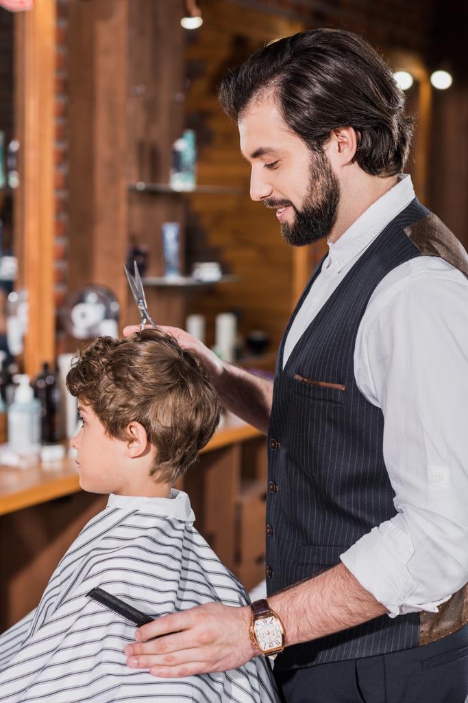 side view of little curly kid covered with striped cloth sitting at barbershop while barber cutting his hair - Photo, Image