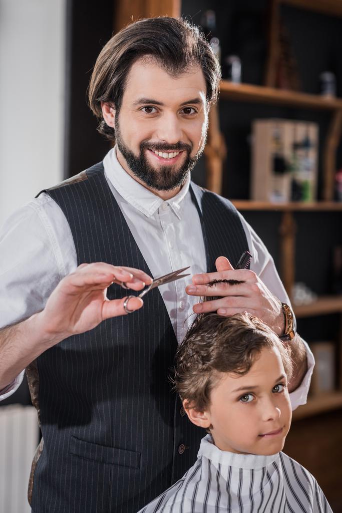 adorable little kid getting haircut from handsome smiling barber at kids barbershop - Photo, Image