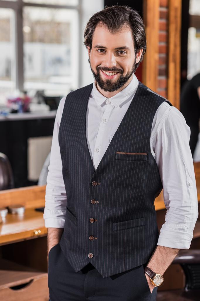 guapo joven peluquero en elegante chaleco mirando a la cámara en la barbería
 - Foto, Imagen