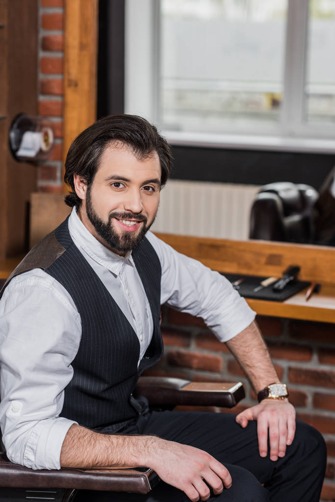handsome barber sitting on chair at workplace and looking at camera - Photo, Image