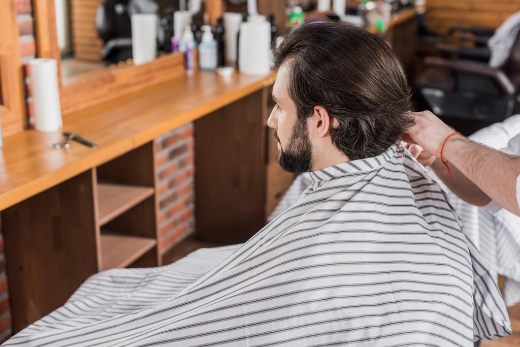 bearded young man covered with striped cloth sitting on chair at barbershop - Photo, Image