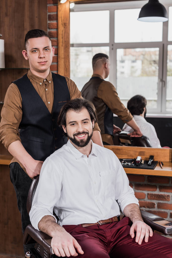heureux jeune client et coiffeur dans salon de coiffure regardant caméra
 - Photo, image