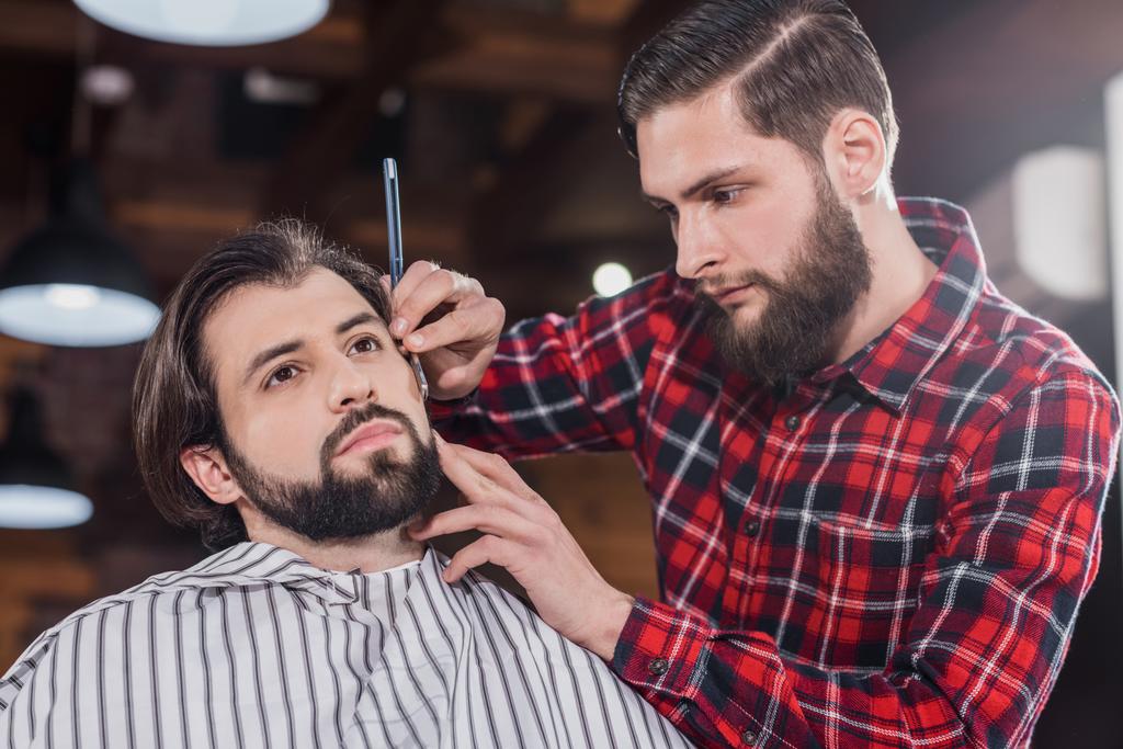 magabiztos fiatal fodrász kockás ing borotválkozás férfi haj vágás géppel - Fotó, kép