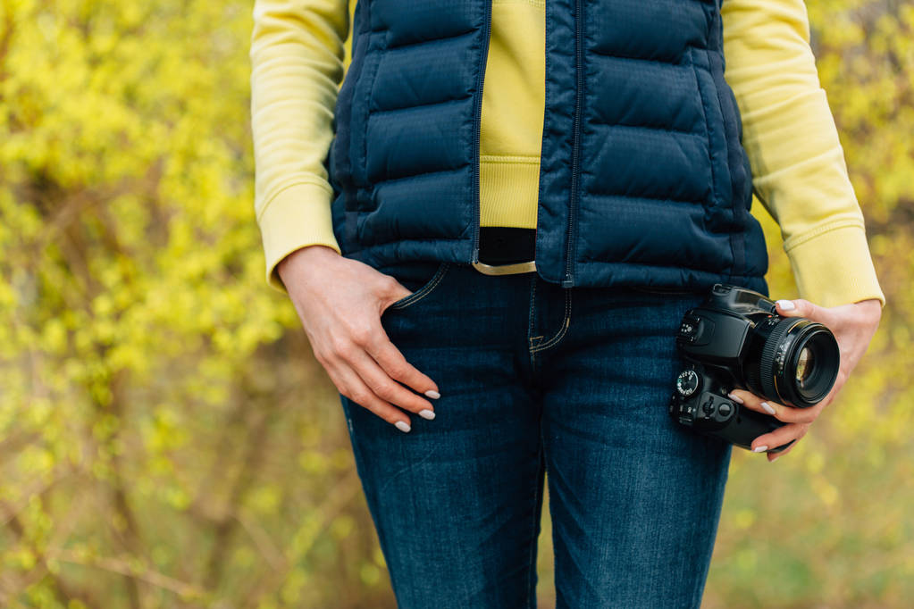 Girl with professional camera in hands standing near forsythia bush. Close up of female photographer in park. - Photo, Image