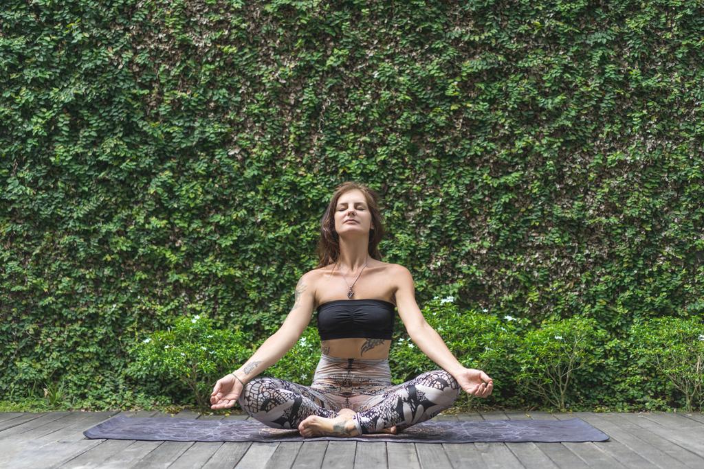 meditando mujer joven practicando yoga en pose de loto frente a la pared cubierta de hojas verdes
 - Foto, imagen