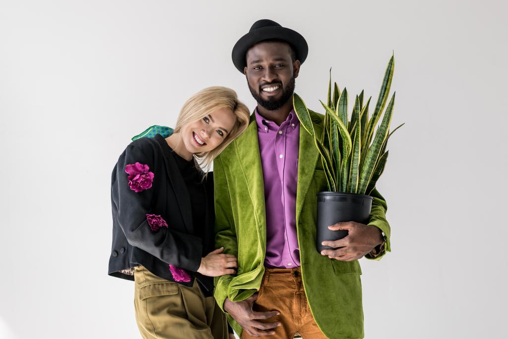 Porträt eines lächelnden gemischtrassigen stilvollen Paares mit grüner Pflanze im Blumentopf, das isoliert auf grau posiert - Foto, Bild