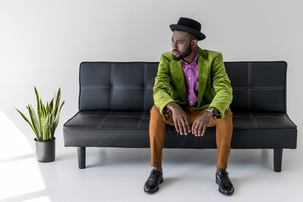 hombre de moda afroamericano pensativo en sombrero sentado en sofá negro
 - Foto, Imagen