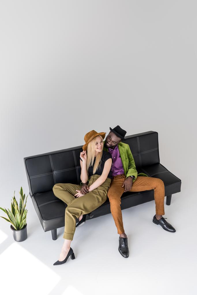 высокий угол обзора улыбающейся мультикультурной модной пары, сидящей на черном диване
  - Фото, изображение