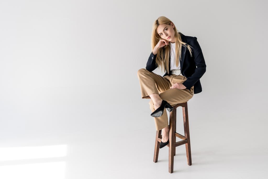houkutteleva nuori blondi nainen tyylikkäissä vaatteissa istuu jakkaralla ja katselee kameraa harmaalla
 - Valokuva, kuva