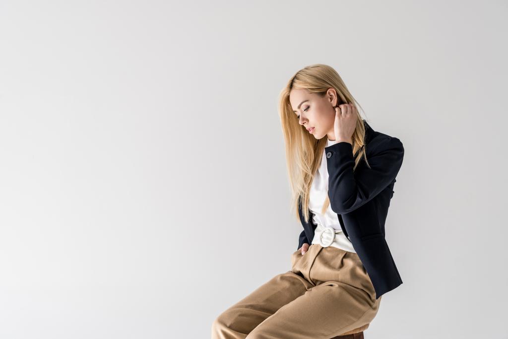 πορτρέτο του όμορφη νεαρή ξανθιά γυναίκα με μοντέρνα ρούχα που κάθεται και κοιτάζοντας προς τα κάτω που απομονώνονται σε γκρι  - Φωτογραφία, εικόνα