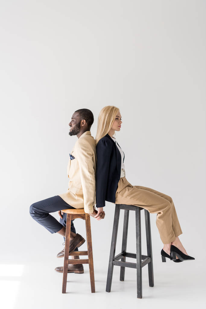 Seitenansicht eines modischen jungen multiethnischen Paares, das Rücken an Rücken auf Hockern sitzt und die Hände auf grau hält - Foto, Bild