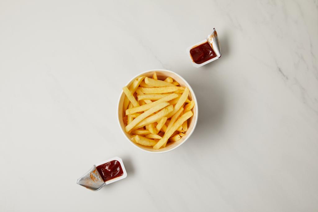 vista superior de papas fritas en tazón con recipientes de ketchup sobre blanco
 - Foto, imagen