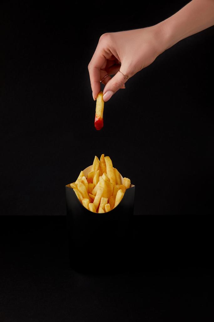 обрезанный снимок женщины, складывающей картошку фри, налитой в кетчуп, поверх коробки картошки фри, изолированной на черном
 - Фото, изображение