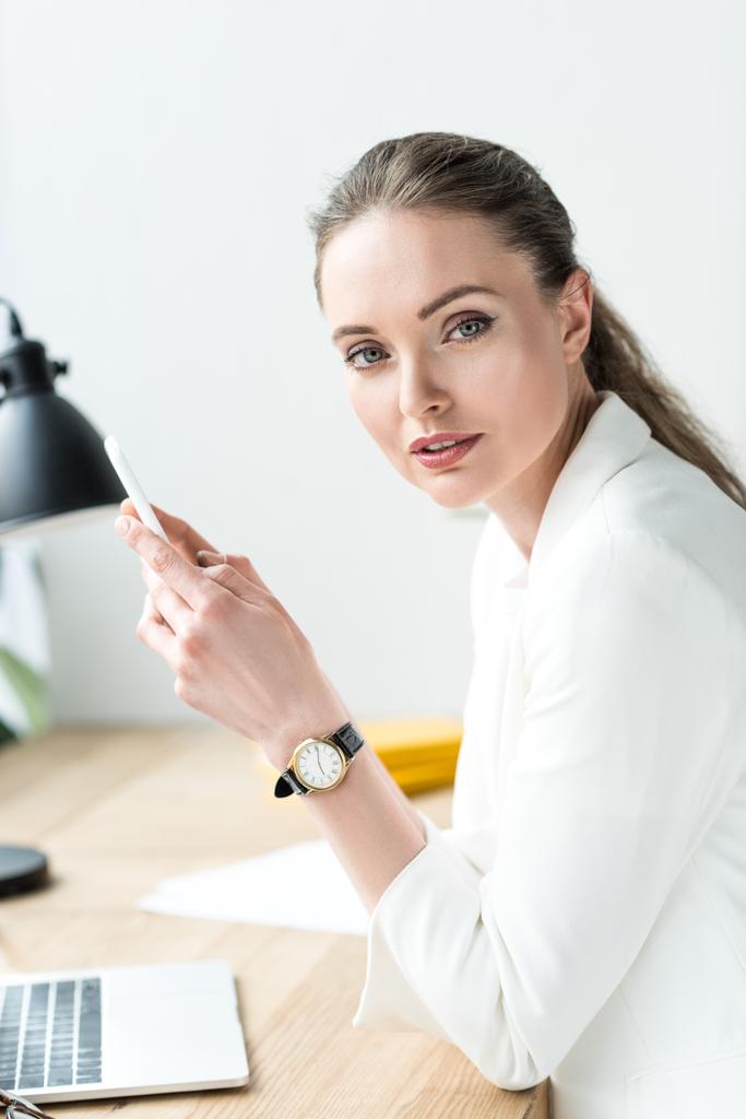 side view of businesswoman in stylish suit with smartphone at workplace with laptop in office - Photo, Image