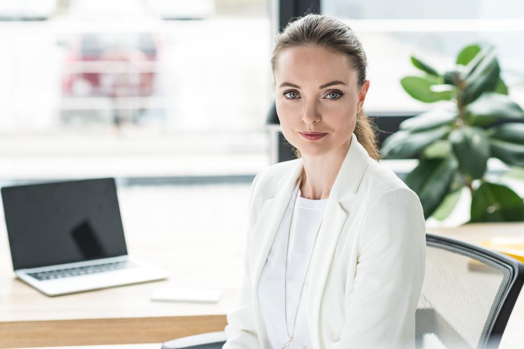 Portret van lachende mooie zakenvrouw in wit pak op werkplek met laptop in office - Foto, afbeelding