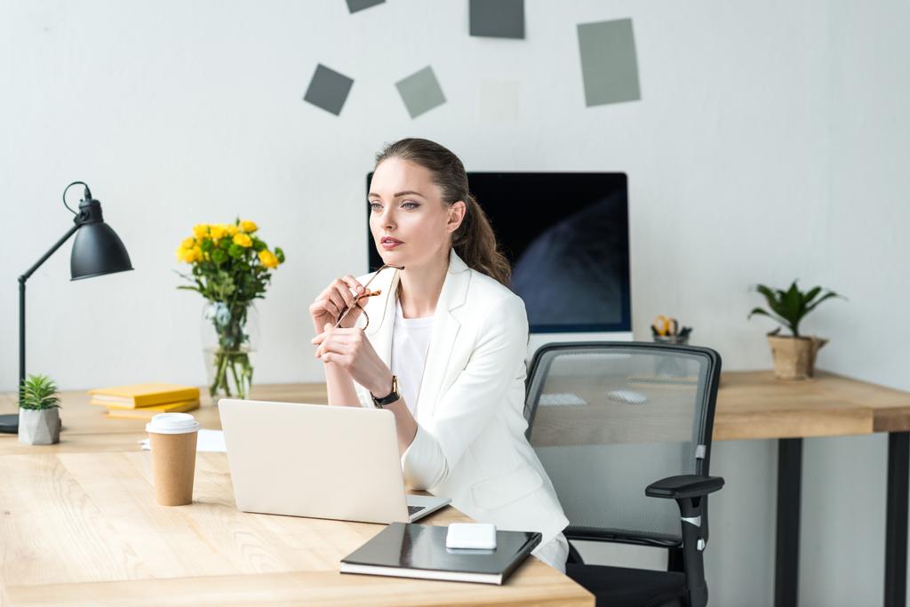 beautiful pensive businesswoman in white suit looking away at workplace with laptop in office - Photo, Image