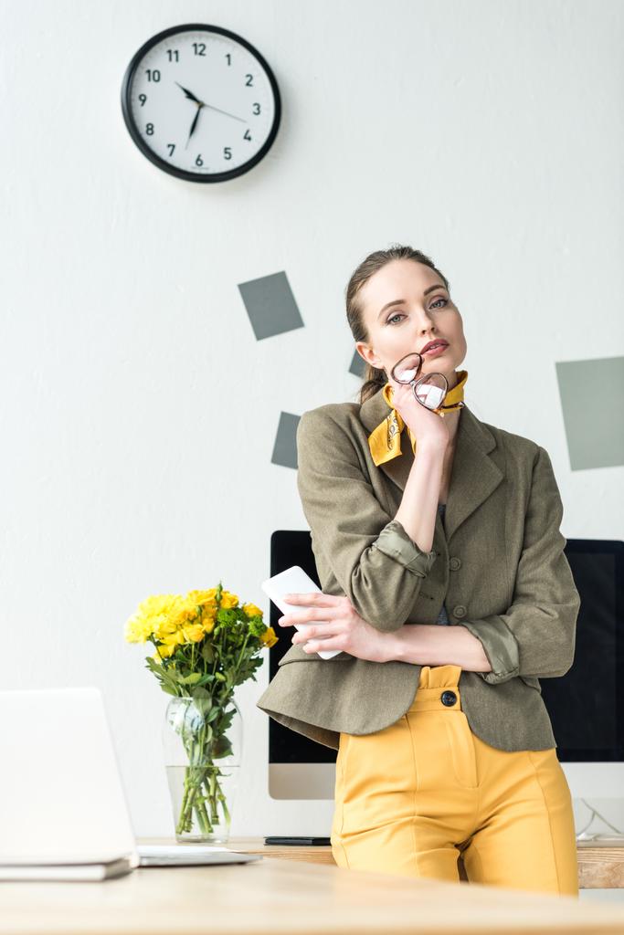 attraente donna d'affari elegante con occhiali e smartphone, guardando la fotocamera in ufficio
 - Foto, immagini