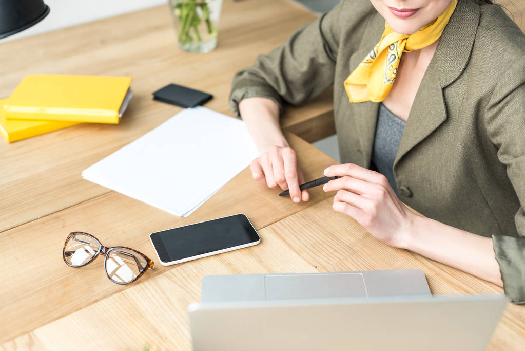 обрезанный снимок стильной деловой женщины, держащей ручку и смотрящей на ноутбук в офисе
 - Фото, изображение