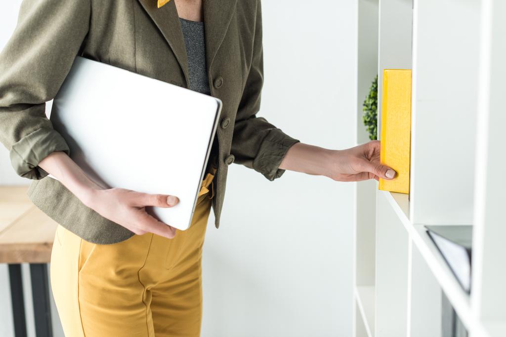 обрезанный снимок предпринимательницы, держащей ноутбук и берущей книгу с полки в офисе
 - Фото, изображение