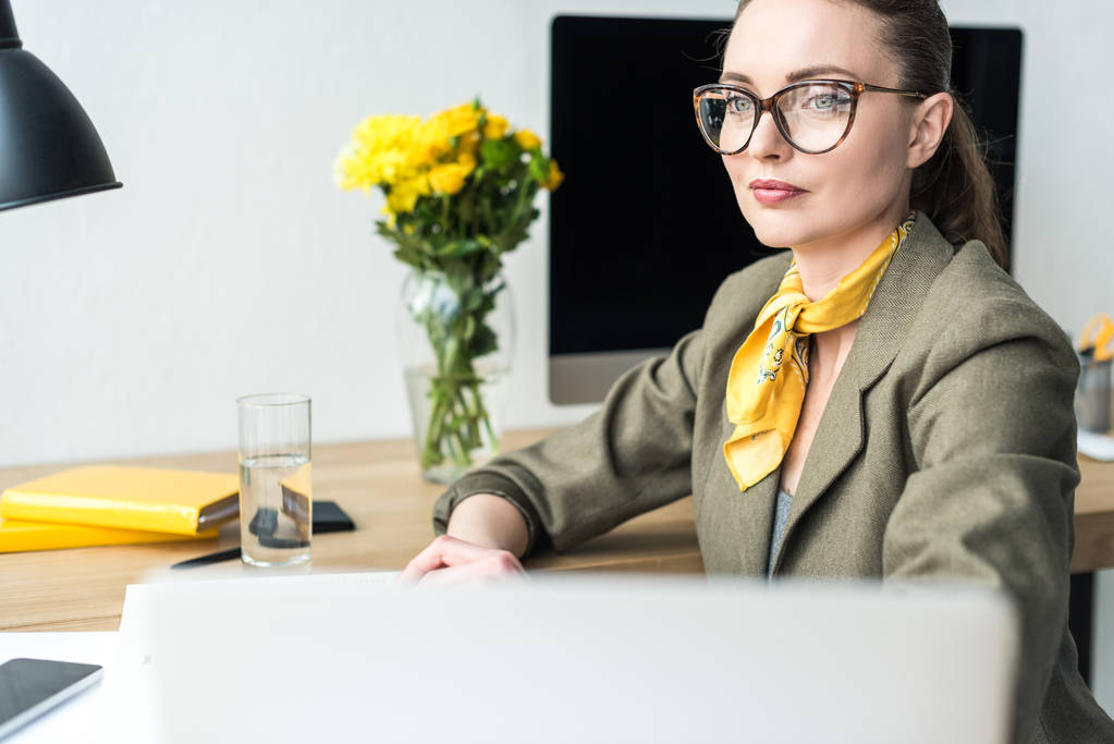 attractive smiling businesswoman in eyeglasses looking away while sitting at workplace - Photo, Image
